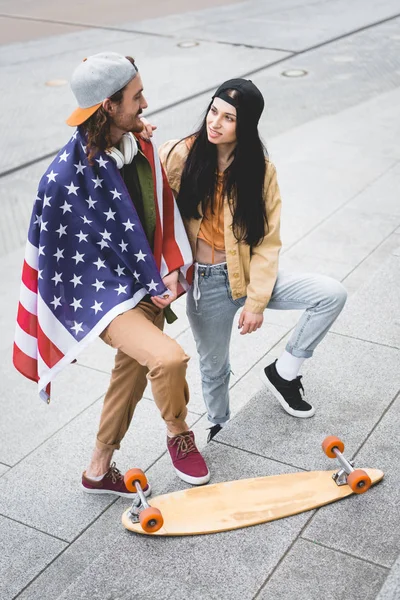 Високий кут зору людини з американським прапором на плечах, дивлячись на брюнетку, що стоїть біля скейтборду — стокове фото