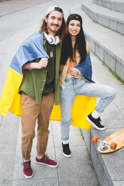Couple heureux avec drapeau ukrainien sur les épaules debout sur les escaliers près de planche à roulettes, regardant la caméra — Photo de stock
