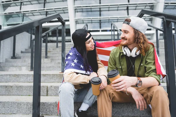 Couple with american flag on shoulders and paper cup in hand sitting on stairs, looking at each other — Stock Photo