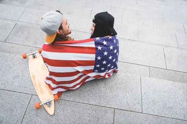 Vista de alto ângulo de casal com bandeira americana em ombros sentados em escadas perto de skate, olhando um para o outro — Fotografia de Stock