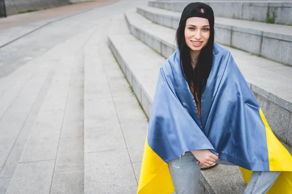 Glückliche Frau mit ukrainischer Fahne auf den Schultern, die auf der Treppe sitzt und in die Kamera blickt — Stockfoto