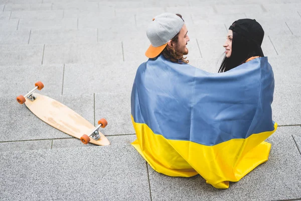 Vista de ángulo alto de la pareja con bandera ucraniana sentado en las escaleras cerca de monopatín, mirándose entre sí - foto de stock