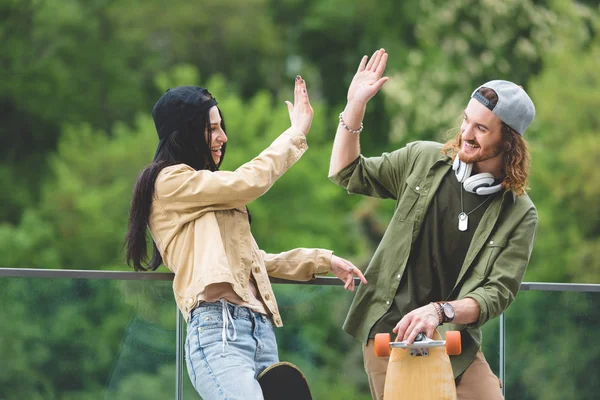 Happy man with skateboard high five to brunette woman, standing on green trees background — Stock Photo