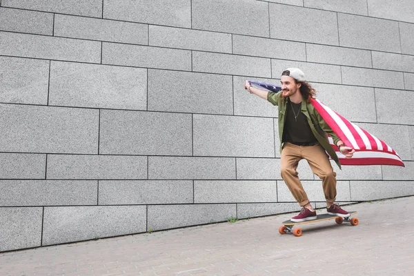 Homem feliz no desgaste casual com bandeira americana montando no skate — Fotografia de Stock