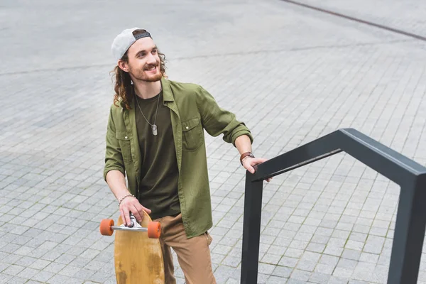 Blick aus der Vogelperspektive auf einen glücklichen Mann in Freizeitkleidung mit Skateboard, der auf der Straße steht und wegschaut — Stockfoto