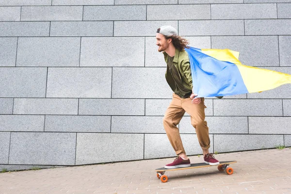 Homme heureux en tenue décontractée et drapeau ukrainien chevauchant sur planche à roulettes — Photo de stock