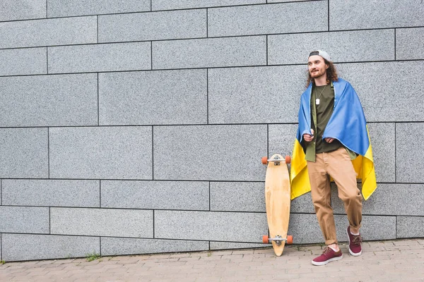 Щаслива людина в українському прапорі на плечах дивиться, стоячи біля стіни з скейтбордом — стокове фото