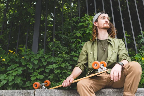 Low angle view of handsome man sitting with skateboard on green plant background, looking away — Stock Photo