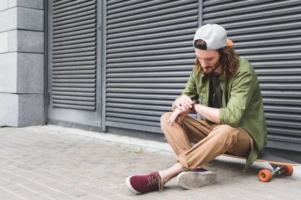 Handsome man sitting on skateboard, looking at smartwatch — Stock Photo