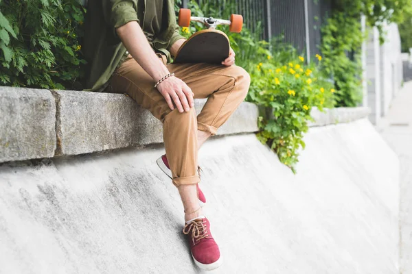 Cropped view of man in casual wear holding skateboard in hand, sitting on border — Stock Photo