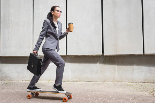 Calm businesswoman in formal wear riding on skateboard, holding paper cup and briefcase in hands — Stock Photo