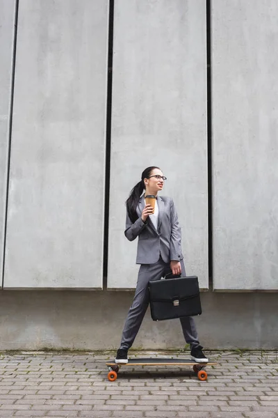 Brunette businesswoman in formal wear riding on skateboard, holding paper cup and briefcase in hands — Stock Photo