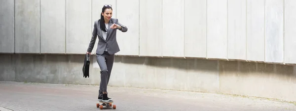 Panoramic view of businesswoman in formal wear riding on skateboard, looking at smartwatch — Stock Photo