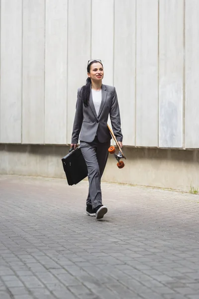Felice donna d'affari in abito formale che cammina per strada con lo skateboard in mano — Foto stock
