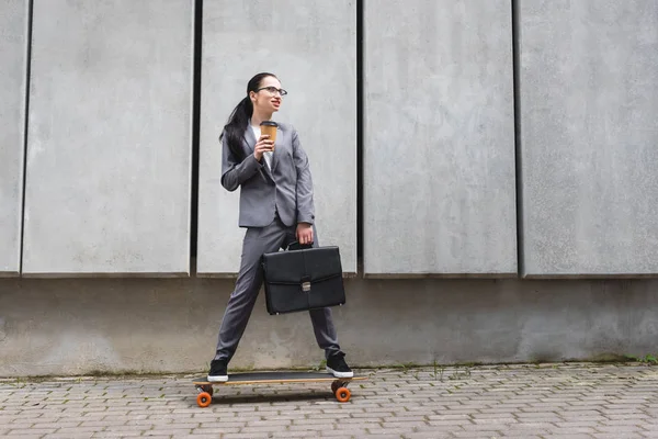 Весела бізнес-леді в офіційному одязі їде на скейтборді, тримає паперовий стаканчик і портфель в руках — стокове фото