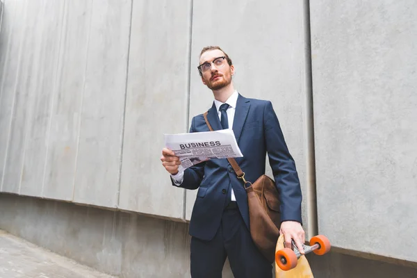 Low angle view of businessman in formal wear standing near skateboard, holding newspaper in hand — Stock Photo