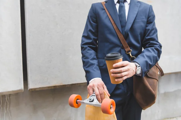 Обрезанный вид бизнесмена в формальной одежде стоя со скейтбордом и бумажной чашкой в руках — стоковое фото
