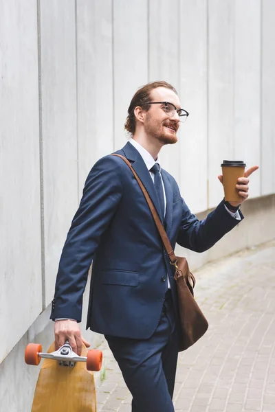 Homem de negócios feliz no desgaste formal segurando skate com copo de papel na mão e apontando dedo longe — Stock Photo