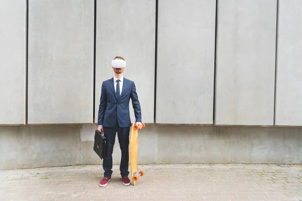 Uomo d'affari in abbigliamento formale e auricolare vr in piedi con skateboard in mano — Foto stock