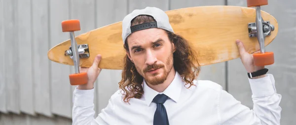 Panoramic view of handsome businessman standing with skateboard, looking at camera — Stock Photo