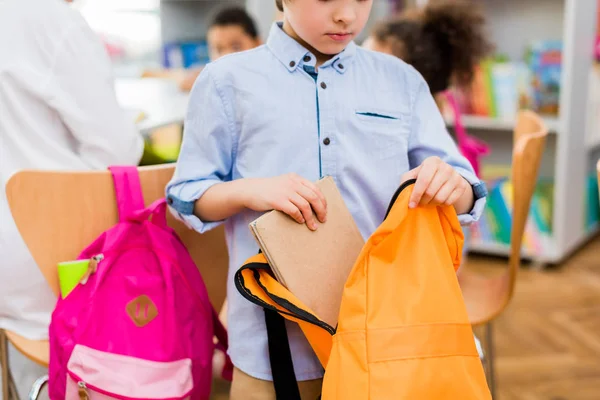 Vista recortada de niño de pie y poner libro en la mochila - foto de stock