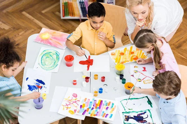 Vue aérienne de la femme regardant des enfants multiculturels peindre sur des papiers — Photo de stock
