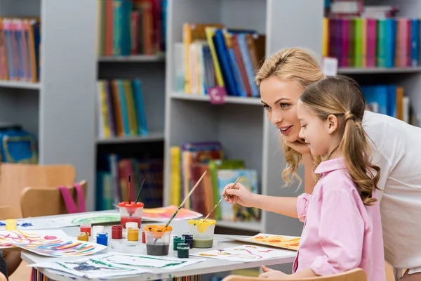 Attraente insegnante vicino carino bambino pittura su carta in biblioteca — Foto stock