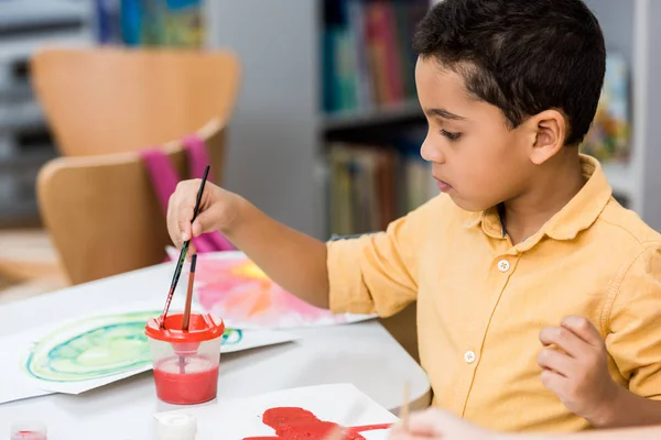 Selective focus of cute african american kid holding paintbrushes near papers with painting — Stock Photo