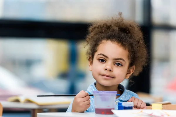 Selektiver Fokus des fröhlichen afrikanisch-amerikanischen Kindes, das in die Kamera blickt und den Pinsel in der Hand hält — Stockfoto