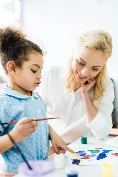 Enfoque selectivo de niño afroamericano apuntando con el dedo a la pintura cerca de la mujer feliz - foto de stock