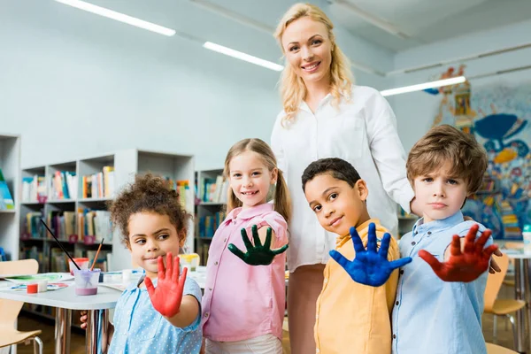 Selective focus of cheerful woman standing with multicultural kids showing colorful hands with gouache paint — Stock Photo
