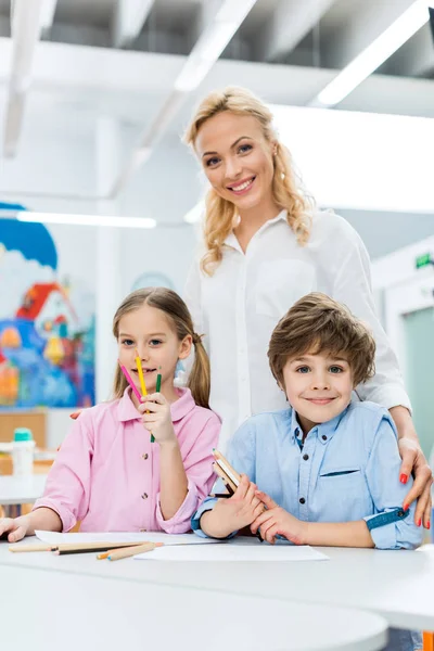 Foyer sélectif des enfants mignons tenant des crayons de couleur et assis près de femme heureuse — Photo de stock