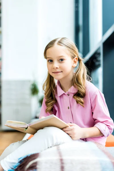 Selektiver Fokus des niedlichen und fröhlichen Kindes, das in der Bibliothek sitzt und Buch hält — Stockfoto