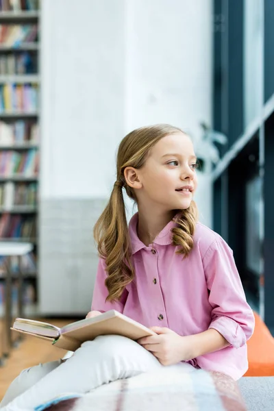 Mignon et coûteux enfant assis et tenant livre dans la bibliothèque — Photo de stock