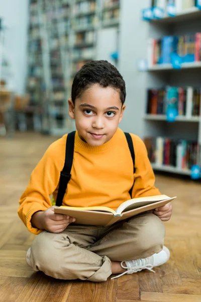 Alegre menino americano africano sentado no chão e segurando livro — Fotografia de Stock