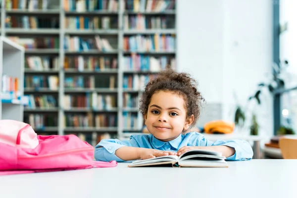 Щасливий афроамериканець дитина сидить і дивиться на камеру біля книги і рожевий рюкзак — стокове фото