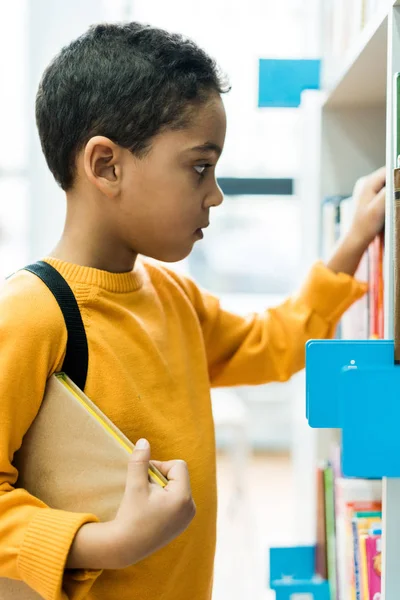 Entzückender afrikanischer amerikanischer Junge steht und hält Buch in Bibliothek — Stockfoto