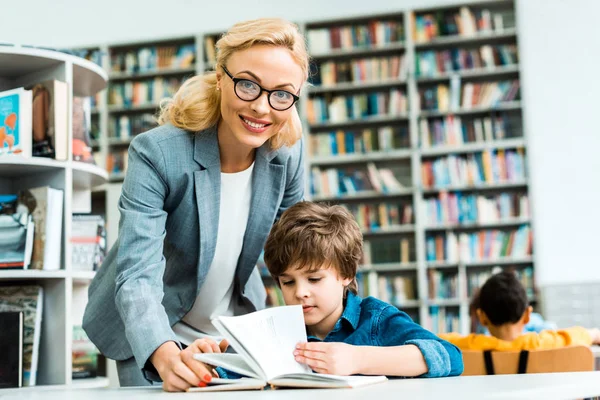 Cheerful woman standing near cute kid reading book in library — Stock Photo