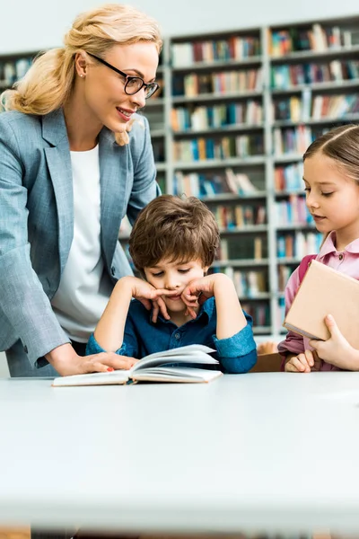 Красивая учительница в очках, стоящая рядом с милыми детьми, читающими книгу в библиотеке — стоковое фото