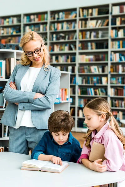 Веселый учитель в очках, стоящий со скрещенными руками и смотрящий на милых детей в библиотеке — стоковое фото