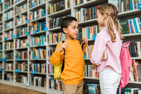 Cute multicultural kids standing and talking in library — Stock Photo