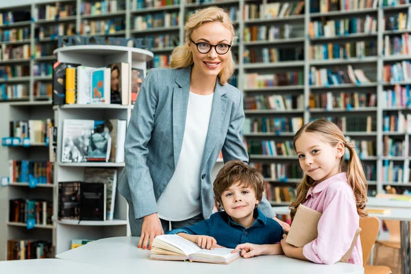 Professeur joyeux dans des lunettes debout près des enfants mignons dans la bibliothèque — Photo de stock