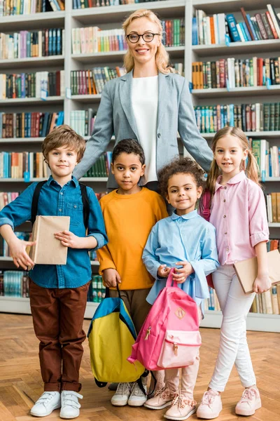 Atractiva y alegre mujer en gafas de pie con lindos niños multiculturales en la biblioteca - foto de stock
