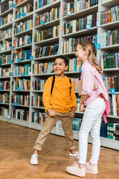 Lindos niños multiculturales de pie y sonriendo en la biblioteca - foto de stock