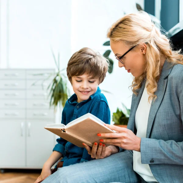 Приваблива жінка в окулярах сидить і читає книгу з щасливою дитиною — стокове фото