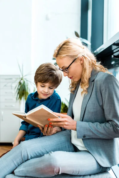Attractive woman in glasses sitting on floor and reading book with cute kid — Stock Photo