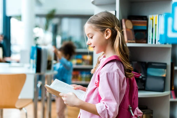 Cheerful kid with pink backpack standing and reading book in library — Stock Photo