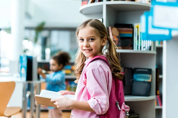 Fröhliches Kind mit rosa Rucksack steht und hält Buch in Bibliothek — Stockfoto