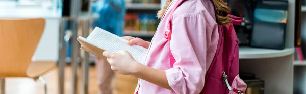 Scatto panoramico di bimbo con zaino rosa in piedi e libro in mano in biblioteca — Foto stock