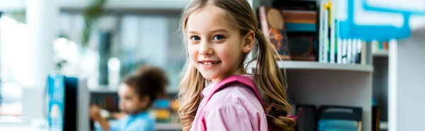 Panoramic shot of cheerful kid with pink backpack standing in library — Stock Photo
